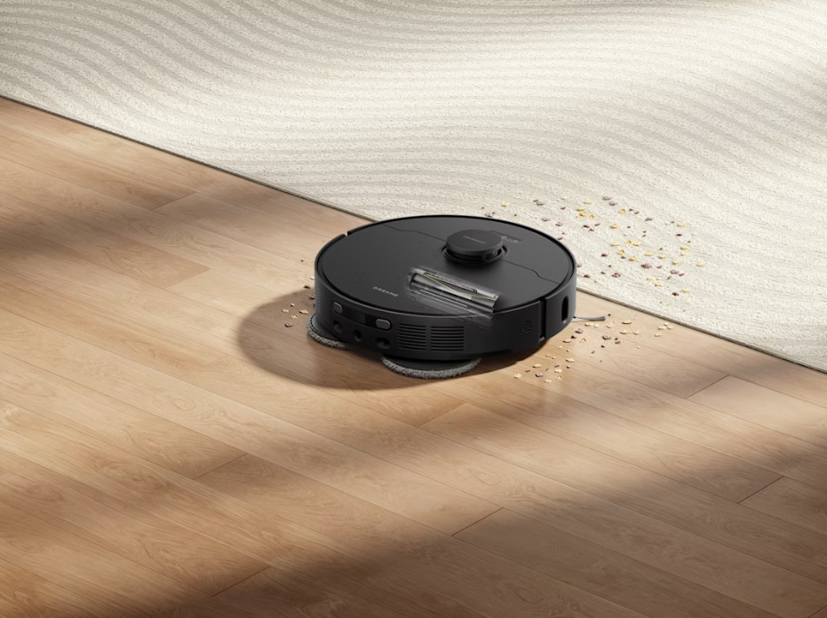 Black MOVA robot vacuum on a wooden floor beside a cream rug scattered with crumbs and pet debris