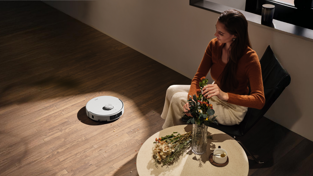 Woman sitting on the floor beside a low table as a white robot vacuum cleans the wooden floor behind her