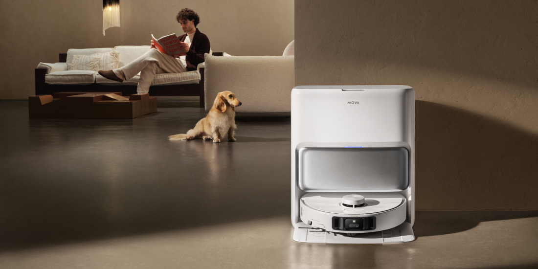 MOVA robot vacuum parked in its self-emptying dock as a dog rests nearby and a person reads on the sofa