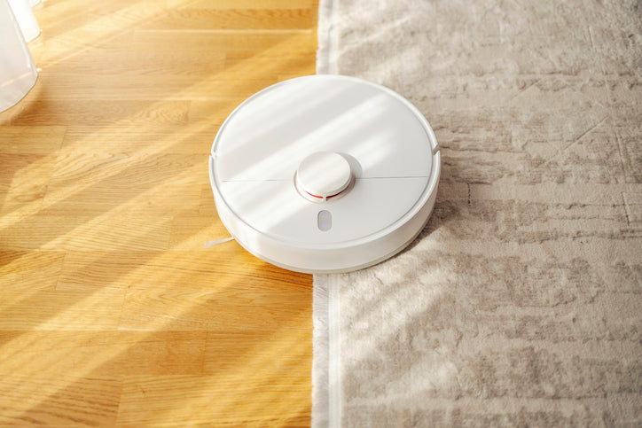 White robot vacuum crossing from a wooden floor onto the edge of a light beige fitted carpet