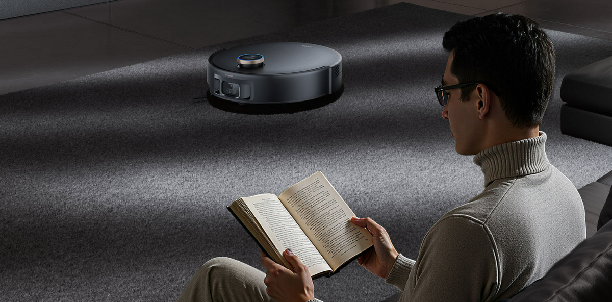 Man reading a book on a dark sofa while a black robot vacuum quietly cleans the carpet behind him